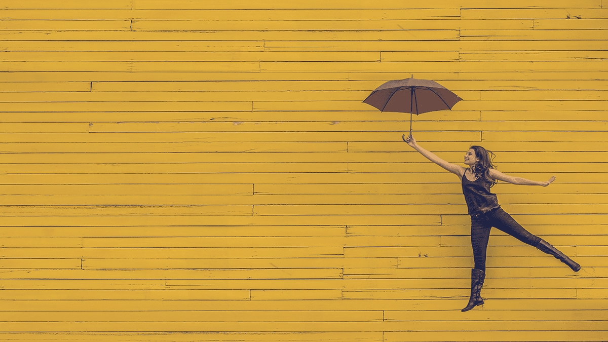 Woman jumping in the air holding an umbrella, in front of a wall of yellow-painted wooden slats.